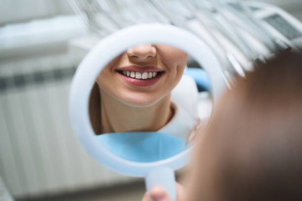 Mujer joven observado sus dientes a través de un espejo. Imagen representativa del post realizado por la clínica dental Avilés Digital Dental Clinic sobre los beneficios en la estética dental de las carillas dentales.
