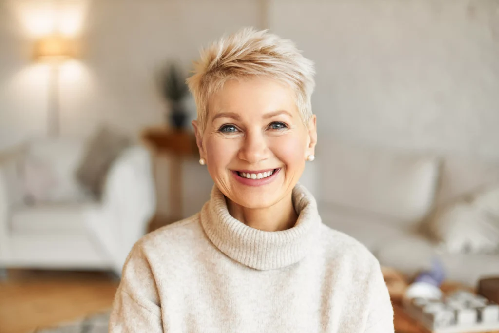 Mujer mayor sonriente con cabello corto y rubio, vistiendo un suéter de cuello alto color crema, en un interior acogedor que refleja un estilo de vida saludable, después del tratamiento de diseño de sonrisa digital.