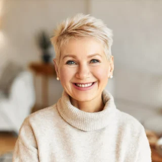 Mujer mayor sonriente con cabello corto y rubio, vistiendo un suéter de cuello alto color crema, en un interior acogedor que refleja un estilo de vida saludable, después de seguir los consejos dentales.