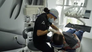 n dentista con uniforme negro y gorro quirúrgico está realizando un tratamiento de blanqueamiento dental en un paciente que está recostado en una silla de dentista. La habitación parece ser un consultorio dental moderno y bien iluminado.