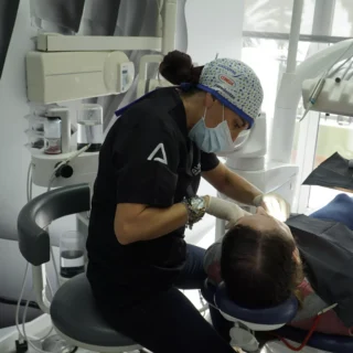 n dentista con uniforme negro y gorro quirúrgico está realizando un tratamiento de blanqueamiento dental en un paciente que está recostado en una silla de dentista. La habitación parece ser un consultorio dental moderno y bien iluminado.
