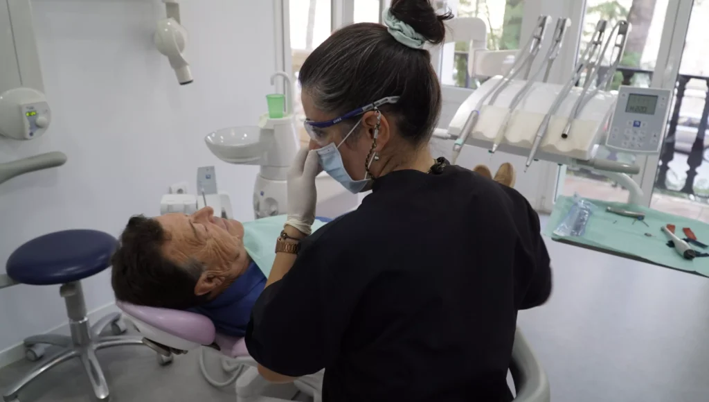 Dentista con atuendo profesional negro revisando a un paciente masculino reclinado en una silla dental en la clínica dental Avilés Digital Dental Clinic, iluminada y moderna.