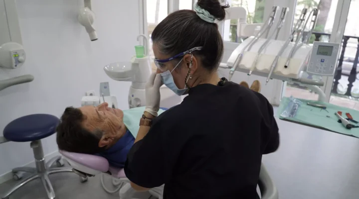 Dentista con atuendo profesional negro revisando a un paciente masculino reclinado en una silla dental en la clínica dental Avilés Digital Dental Clinic, iluminada y moderna.